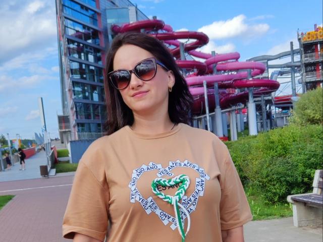 Izobella69 met donkerbruin haar, die een zonnebril draagt, een beige shirt met een groene slang erop, staat buiten en toont een zelfverzekerde glimlach. Ze heeft kleine borsten en een schattige achterkant..