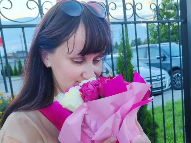 Izobella69, avec des cheveux bruns, des lunettes de soleil sur la tête, souriante, tenant un bouquet de roses roses, un ruban rose, une rose blanche, du papier rose, à l'extérieur, une clôture en fer noir, une voiture en arrière-plan, de l'herbe..