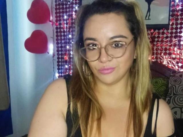 NaugthyCurvy, avec des lunettes, du rouge à lèvres rose et des cheveux bruns, portant un haut noir, a un visage mignon, légèrement rond, et regarde l'appareil photo avec un léger sourire..