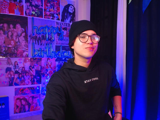 EddTerence with glasses, black beanie, and black hoodie, smiling, standing in front of colorful wall photos, blue and purple lighting, tattoo on right forearm.
