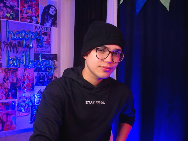 EddTerence, wearing a black beanie and glasses, has a cute face and is dressed in a black Stay Cool hoodie, sitting in a room with blue lighting.