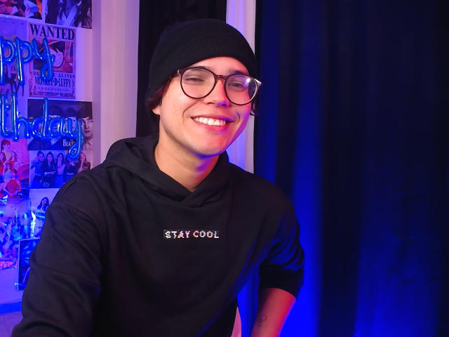 EddTerence, smiling with glasses and black beanie, wearing a black hoodie with "STAY COOL" text, has a cute face and is showing his teeth. Blue and purple light.