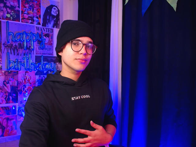 EddTerence is a nerdy dude with glasses, a black beanie, and a black "STATICAL" hoodie, touching his chest, standing in a blue-lit room with birthday decorations.