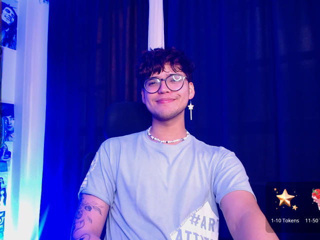 EddTerence has curly brown hair, glasses, and a cross earring. He's wearing a light blue shirt with a white triangle, and a beaded necklace. His arm has a tattoo.