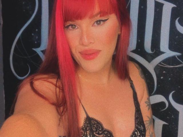 VictoriaFoxys with bright red hair, black lace bra, and tattoos, smiling at the camera, showing her tits, with bold eyeliner and pink lipstick.
