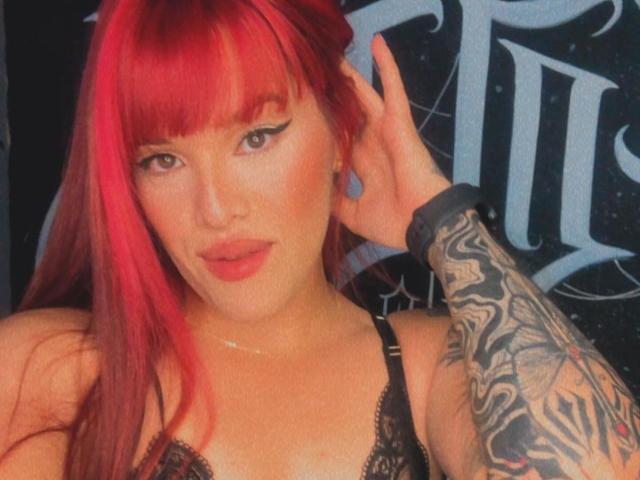 VictoriaFoxys with bright red hair, black eyeliner, and tattoos on her right arm, wearing a black lace bra, touching her ear, and looking at the camera.