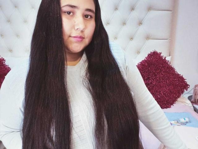 ZoeGiil with long black hair, wearing a white long-sleeve shirt, sitting on a bed with red pillows, looking at the camera with a slight smile.