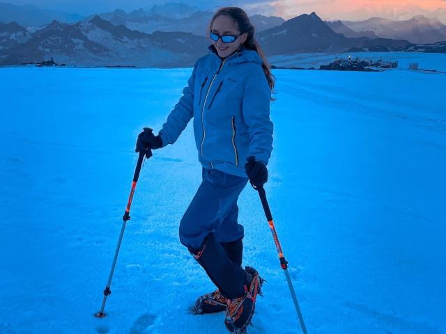 BiancaHotLove with blue ski jacket, black gloves, and sunglasses, holding ski poles, standing in snow, wearing orange ski boots, smiling, in a snowy mountain landscape at dusk.