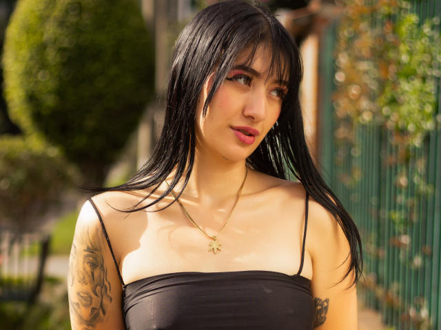 TifanyDiaz with black hair, pink lipstick, and tattoos on her right arm, wearing a black strap top, gold necklace, and hoop earrings, smiling outdoors.
