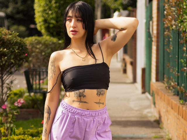 TifanyDiaz with black tube top, showing her small tits, purple pants, and tattoos, standing outside with one arm behind her head, and wet black hair.