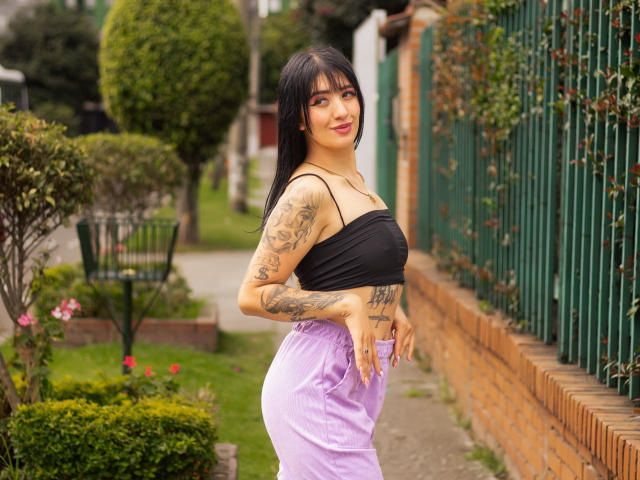 TifanyDiaz with tattoos, black tube top, and purple pants, standing with a slight smile, hands in pockets, black hair, red lipstick, and a curvy, sexy ass.