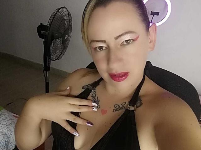 LindaMilejets with tattoos on her chest, wearing a black halter top, purple nails, and red lipstick, holding her tits, fan in background, ring light on wall.