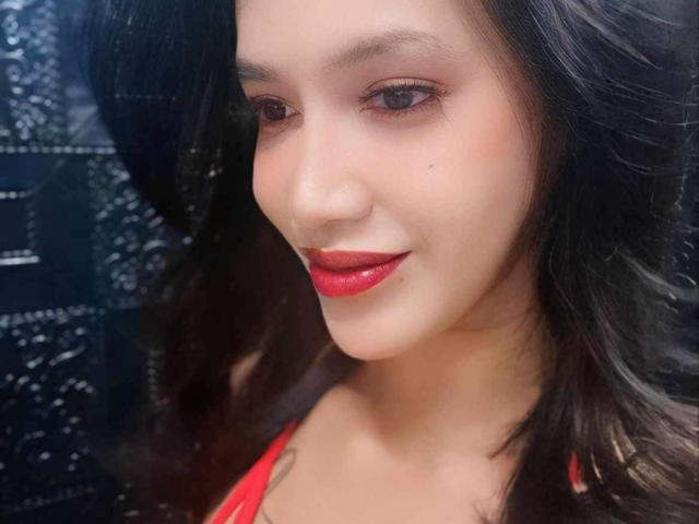 Le visage de CumEaterJanna, rouge à lèvres, léger sourire, cheveux noirs ondulés, peau claire, maquillage discret, une petite tache visible près de l'œil gauche, portant un haut rouge, fond noir texturé..