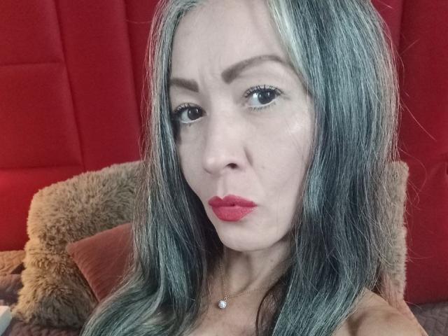 Nicolette Shea tem cabelos longos, prateados e esverdeados, batom vermelho e uma expressão séria. Ela está usando um colar com uma pequena pedra branca e redonda. Seus seios estão parcialmente visíveis..