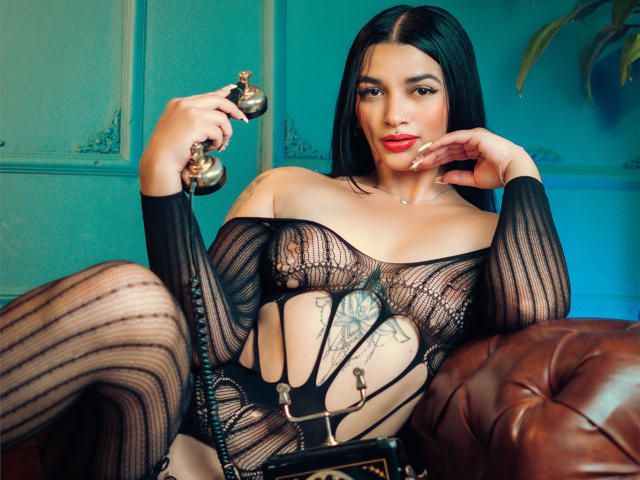 BellaMontez with black hair, wearing sheer black lingerie, holding a vintage phone, sitting on a brown leather couch, showcasing her tits and a tattoo on her lower belly.