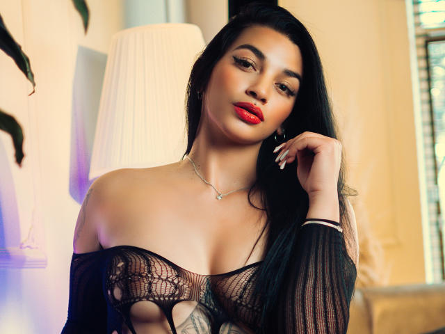 BellaMontez with long black hair, red lipstick, and black sheer top, exposing her tits, hand on cheek, necklace, and earrings, in a softly lit room.