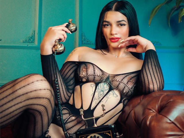 BellaMontez with black mesh lingerie, holding a vintage phone, sitting on a brown leather chair, showing her tits and tattooed belly, with red lipstick and long black hair.