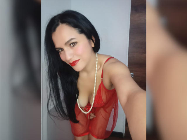 CameronViviane with long black hair, red lace lingerie, pearl necklace, and red lipstick, leaning forward, showing her tits, taking a selfie in a mirror.