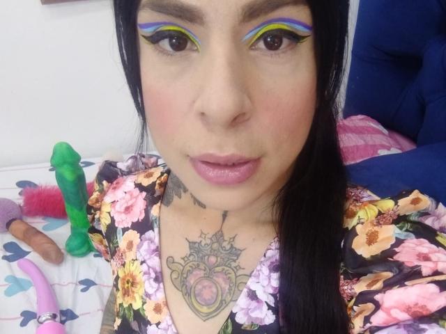 SaoryPrincess with bold blue and yellow eyeliner, pink lips, floral shirt, chest tattoo, black hair, and a green dildo on a bed. Her tits are partially visible.
