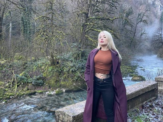 SweetNosha with long blonde hair, wearing a brown crop top, black jeans, and a purple coat, standing by a rocky stream, looking slightly to the side with a relaxed expression.