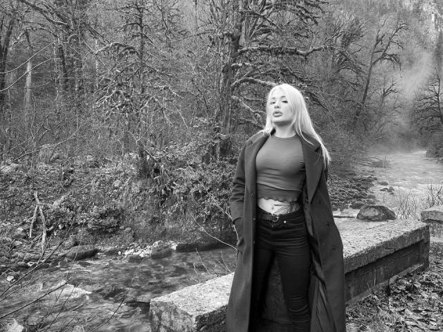 SweetNosha with long blonde hair, wearing a torn crop top, black jeans, and a long coat, standing by a rocky stream, looking slightly to the side with a relaxed expression.