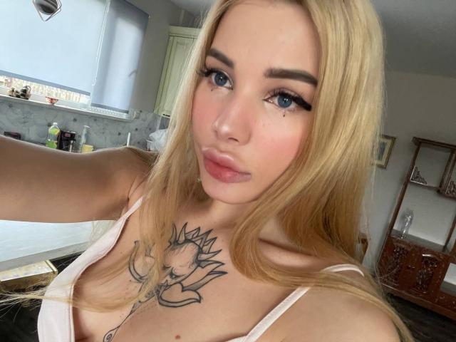 SweetNosha with blonde hair, blue eyes, and pink lipstick, showing off her tattoos, including a sun face on her chest, in a pink top, with her boobs slightly visible.