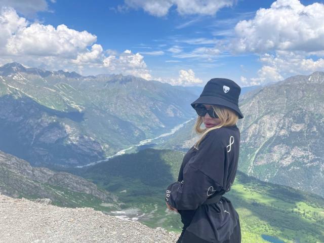 SweetNosha with blonde hair, black hat, sunglasses, and black outfit, showing her ass, standing on a mountain with a valley and mountains in the background.