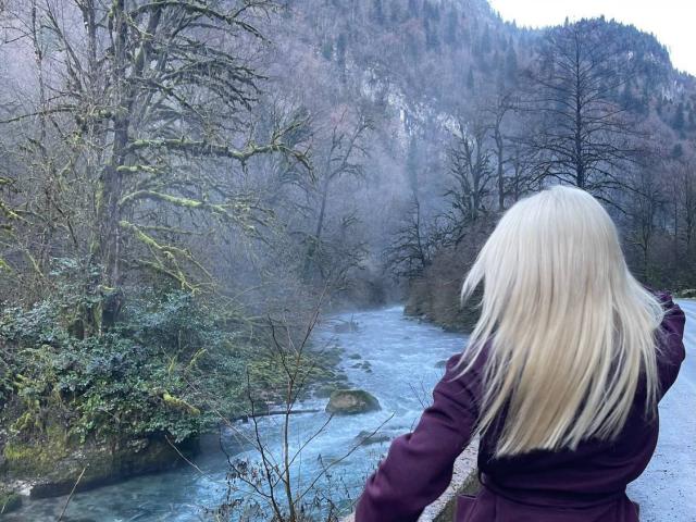 SweetNosha with long blonde hair, facing a blue river, wearing a dark purple coat, standing in a forested mountain area, with trees and rocky hills in the background.