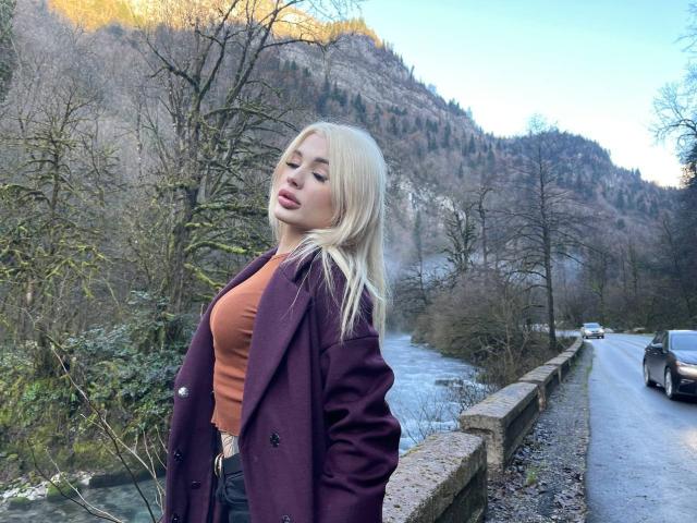 SweetNosha with blonde hair, eyes closed, wearing a purple coat and orange top, standing on a road beside a flowing river, hands in coat pockets, looking relaxed.