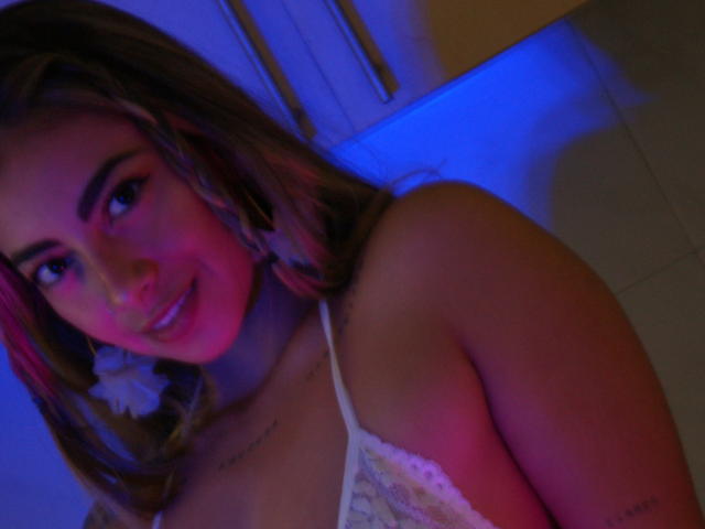 Alessasantini with pink and blue lighting on her face, smiling, wearing white lace lingerie, visible tattoos, and pink-tinted brown hair, looking at the camera.