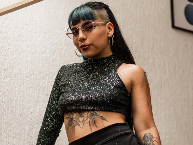 SusanDarknes with teal-streaked black hair, glasses, hoop earrings, and a glittery black crop top, showing a large tattoo on her lower abdomen and a black skirt.