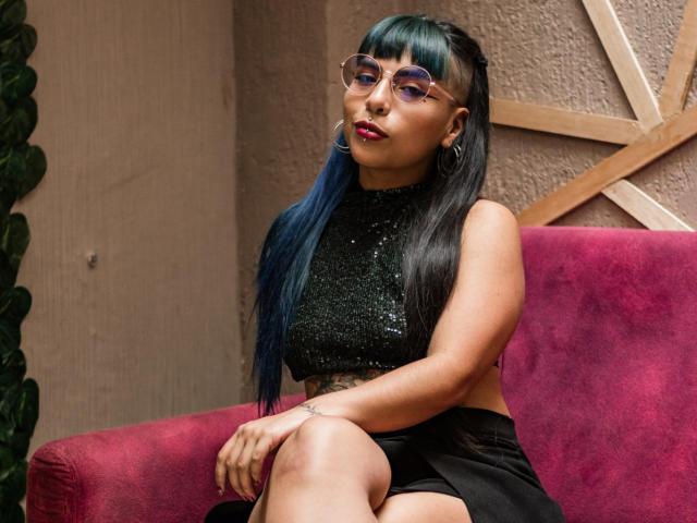 SusanDarknes with blue hair, wearing glasses, a black sequined top, and black skirt, sitting on a pink couch, showing her tattooed arm and crossed legs.