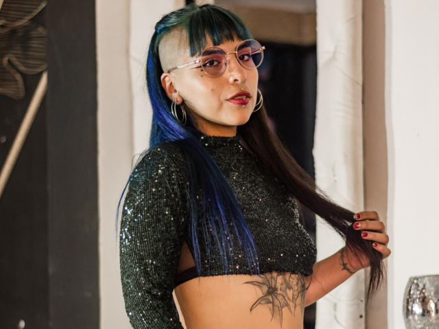 SusanDarknes with blue and black hair, shaved side, glasses, hoop earrings, sparkly black top, holding hair, tattoo on side, pink lipstick, standing, looking at camera.