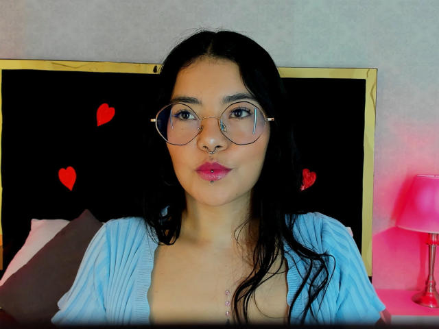 EmmaRuiz with long black hair, round glasses, nose ring, pink lipstick, and a blue cardigan, showing her small tits, sitting against a black headboard with red heart stickers.