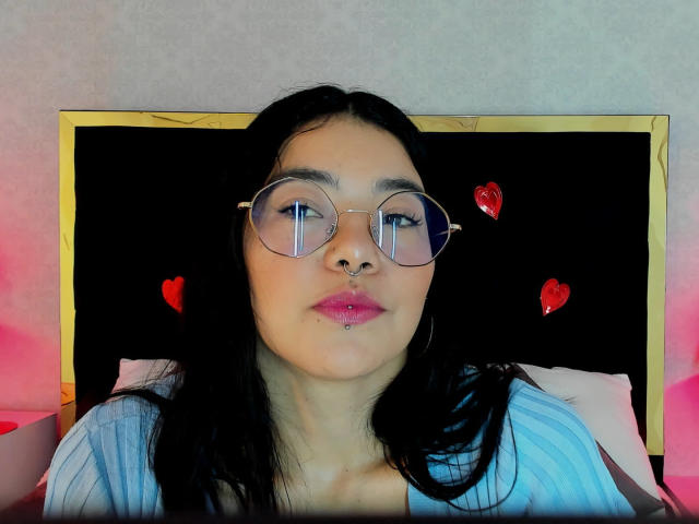 EmmaRuiz with black hair, round glasses, nose ring, lip ring, pink lipstick, wearing a blue shirt, red heart stickers on a black headboard, pink and red lighting.