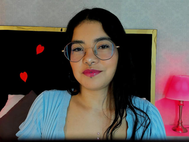 EmmaRuiz with dark hair, glasses, nose ring, pink lipstick, and blue cardigan, showing her tits, sitting in front of a black headboard with red heart stickers.