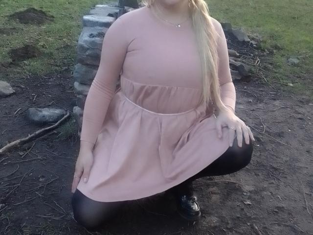 KelliRedhaer squatting, pink dress, black tights, blonde hair, long sleeves, dirt ground, stone wall, black shoes, hands on knees, visible ass, necklace, grassy background.