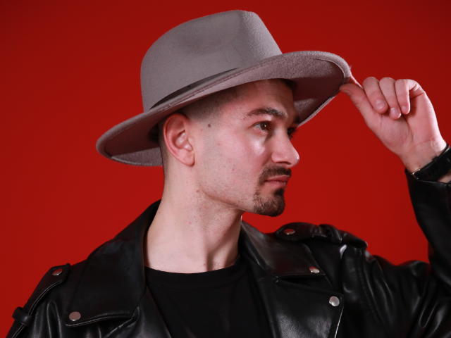 Sergio Bravo trägt einen grauen Hut, eine schwarze Lederjacke und ein schwarzes Hemd. Er justiert seinen Hut mit ernstem Gesichtsausdruck vor einem roten Hintergrund. Er hat einen kurzen Bart..