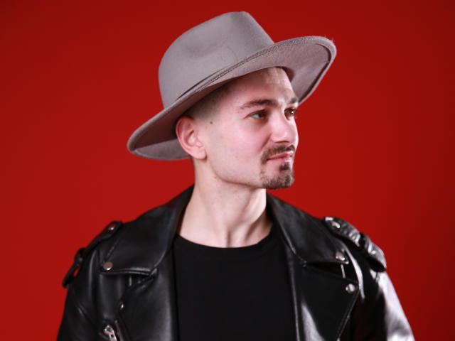 Sergio Bravo mit einem grauen Hut, einem schwarzen Hemd und einer schwarzen Lederjacke, der zur Seite schaut, mit Schnurrbart und Bart, vor einem einfarbigen roten Hintergrund..