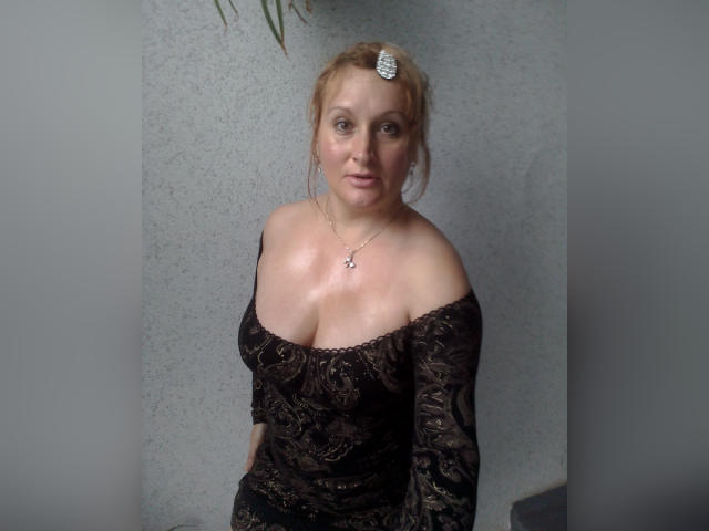 KelliRedhaer has blonde hair, a jeweled hairpin, and a black lace off-shoulder top showing her big boobs and cleavage. She's wearing a silver necklace and looking at the camera.