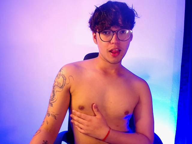 Shirtless EddTerence with glasses, dark curly hair, tattoo on left arm, hand covering small, soft, brown nipples, blue and purple light, red bracelet, open mouth, looking at camera.