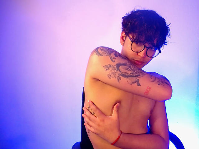 Shirtless EddTerence with tattoos on his arm and chest, wearing glasses, hugging himself with a ring on his hand, a red bracelet, and a sad expression.