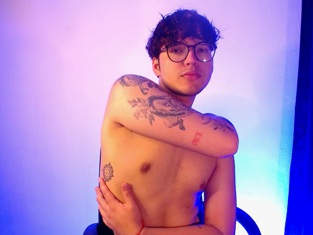 EddTerence, shirtless, with curly brown hair, glasses, and tattoos, crosses arms over chest, looking at camera, showing small tits and a hand on his ass. Blue and purple light.