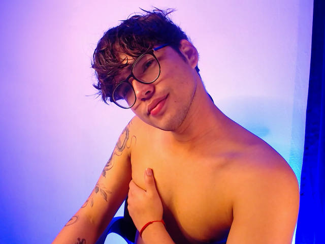 Shirtless EddTerence with curly brown hair, glasses, and tattoos, holding his left shoulder, wearing a red bracelet, and smiling slightly. Blue and purple lighting.