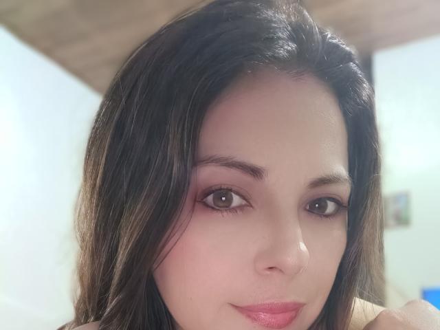 Seher's face, showing brown eyes, pink eyeshadow, and pink lipstick, with straight dark brown hair and a slight smile, in a soft-lit indoor setting.