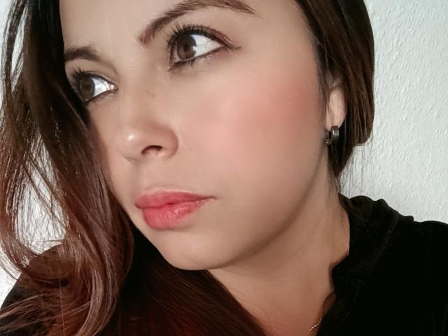 Seher has light brown skin, brown eyes looking to the side, pink lipstick, and brown hair with a slight wave. She wears a black top with small earrings.