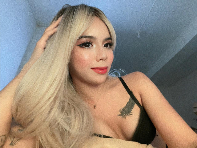 AyeshaSebastian, avec ses longs cheveux blonds, son soutien-gorge noir et un tatouage de feuille sur sa poitrine droite, sourit à l'appareil photo, une main dans ses cheveux. Elle est jolie, avec du rouge à lèvres et un eye-liner..