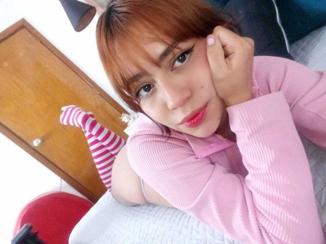 Anni Evans, con i capelli rossi, calze a righe rosa e una maglia a coste rosa, sdraiata sul letto, sorridente, con un viso carino, labbra rosa e una piccola lentiggine sulla guancia..
