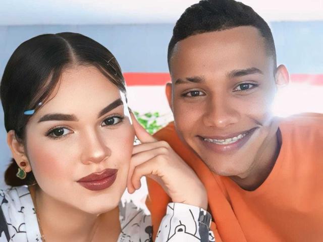 MorenoAndNayla with dark hair, makeup, and maroon lipstick, and Nayla with short black hair, orange shirt, smiling, touching MorenoAndNayla's face. Bright sunlight from right.