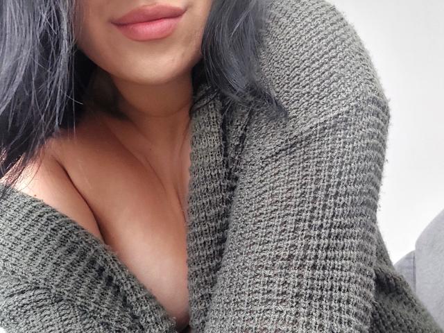 LauraRichis with dark hair, wearing a green knitted sweater, exposing her cleavage and medium-sized tits. Her pink lips are slightly parted. She's in a soft, natural light.